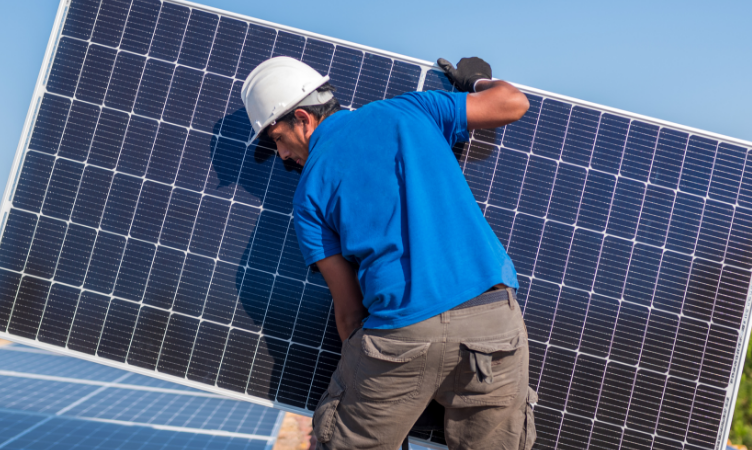 Homem branco de costas usando capacete segurando uma placa solar grande - geração própria de energia