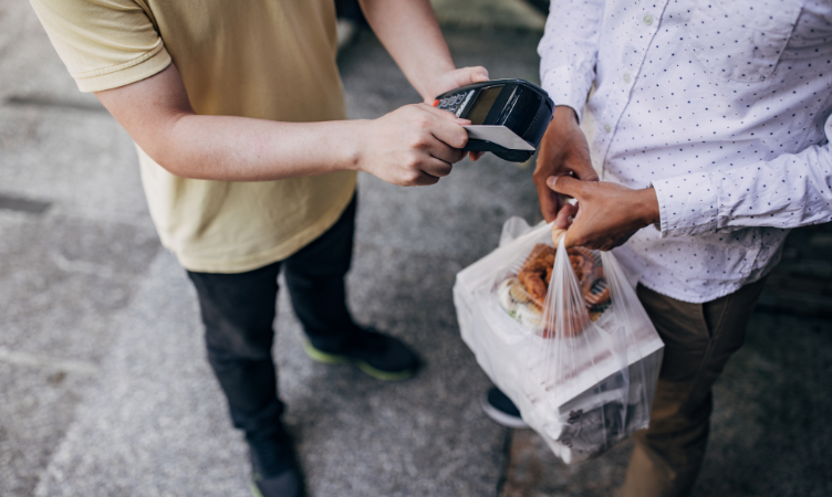 em uma calçada, aparece um homem segurando uma maquininha de cartão e outro homem segurando uma sacola com uma compra
