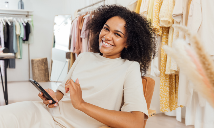 Mulher negra sorrindo sentada em uma cadeira dentro de uma loja de roupas fazendo a gestão de estoque no celular
