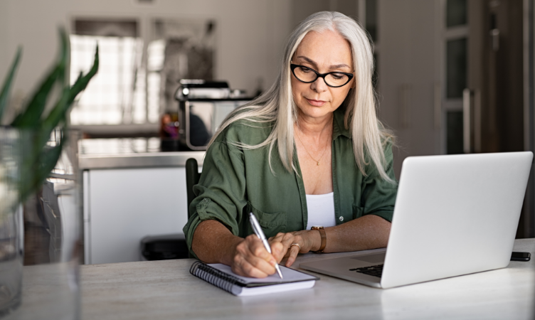 mulher branca de meia-idade, cabelos longos e óculos está apoiada em uma mesa com um notebook, fazendo anotações em um pequeno caderno