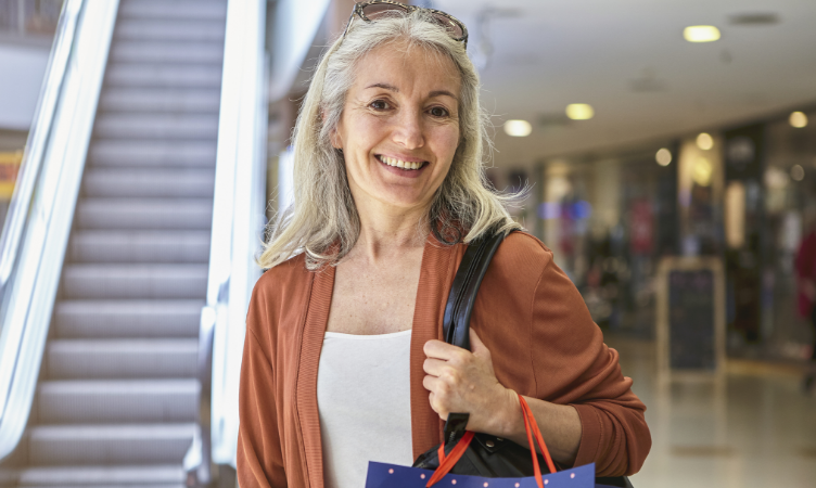Imagem destaque do blogpost do Sebrae RS sobre economia prateada no Brasil. Nela temos a imagem de uma senhora de cabelos brancos e longos sorrindo e segurando sacolas de compras dentro de um shopping. No fundo, aparece uma escada rolante e lojas.