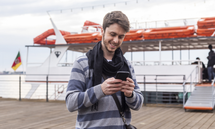 homem jovem sorridente olhando para a tela de um celular, ao fundo tem uma lancha de passeio