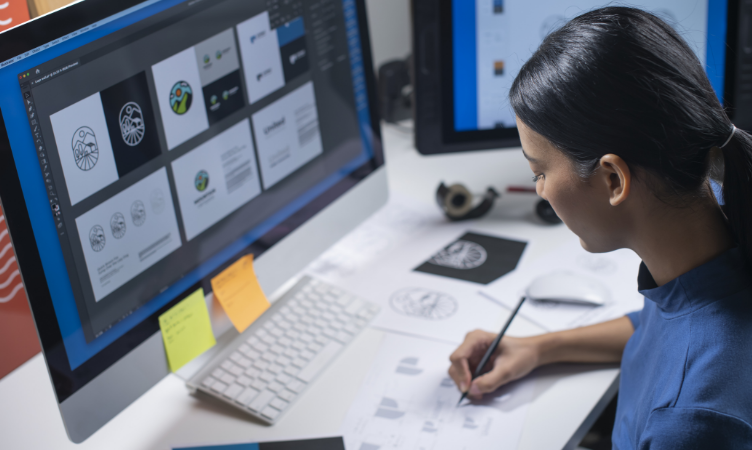 Imagem destaque do blog post do Sebrae RS sobre design para pequenos negócios. Nela, temos a imagem de uma mulher sentada na frente de um iMac fazendo estudos de marca. Ela desenha opções em um papel enquanto olho para a tela.