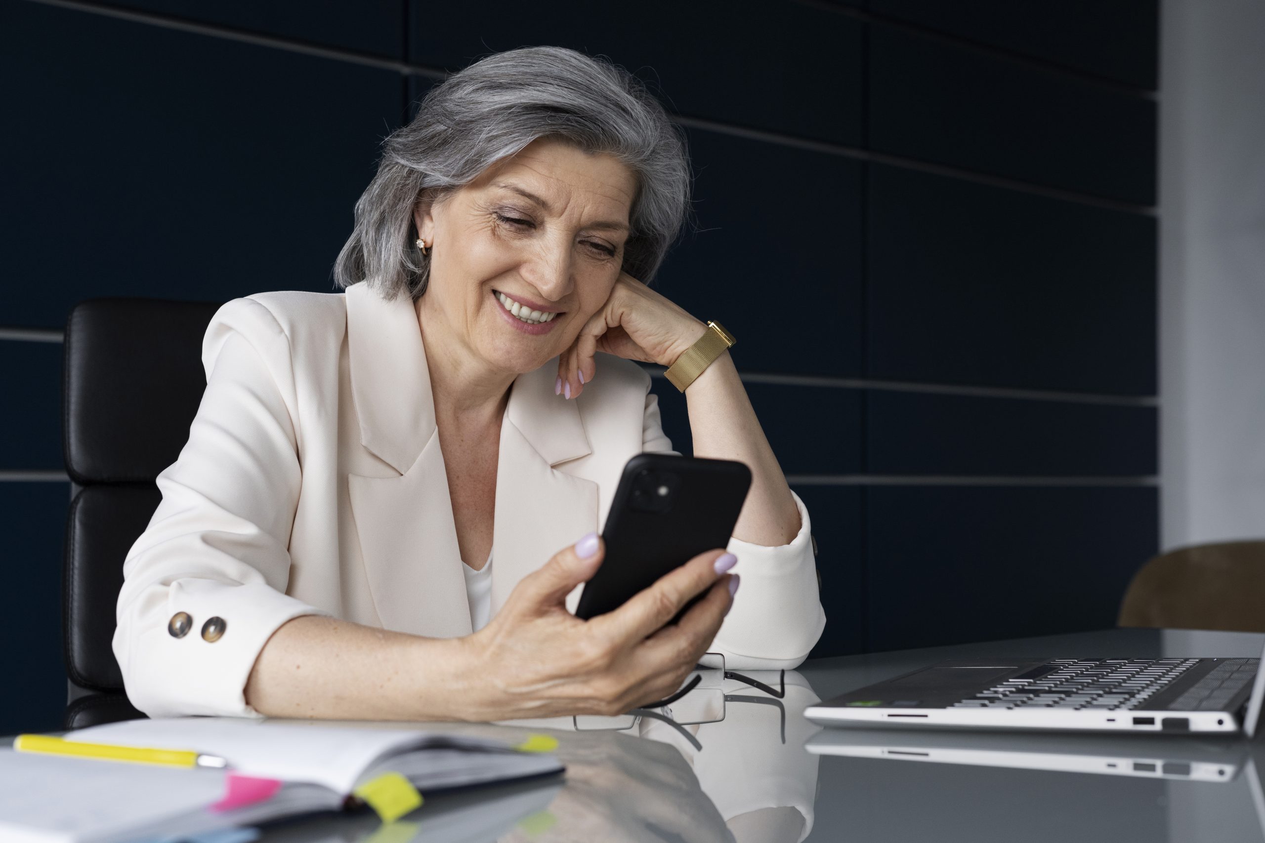 Empreendedorismo 60+ - imagem de mulher idosa, com um blazer branco, sentada em uma mesa, olhando para a tela do celular e sorrindo.