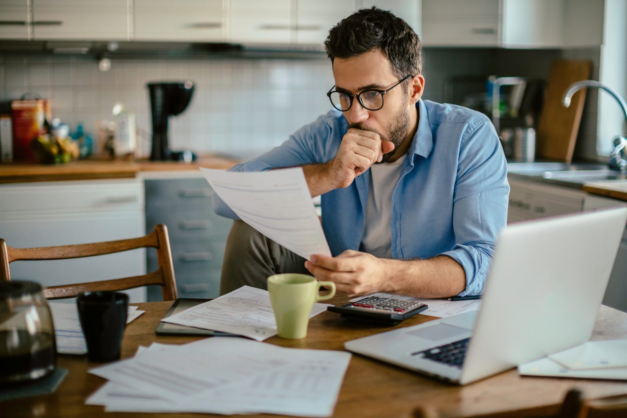 blogpost débitos da dívida ativa - homem sentado a mesa, com notebook, papéis e calculadora, com expressão de aflição olhando para um papel.