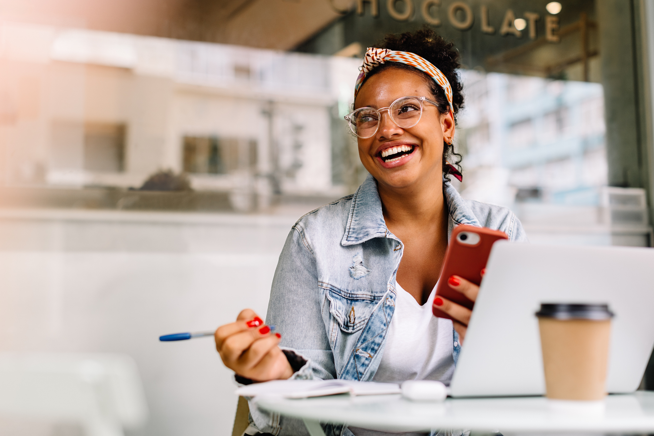 Imagem blog Marketing de Comunidade. Mulher sorrindo em uma cafeteria, utilizando um notebook e um celular enquanto trabalha ou conversa em um ambiente descontraído.