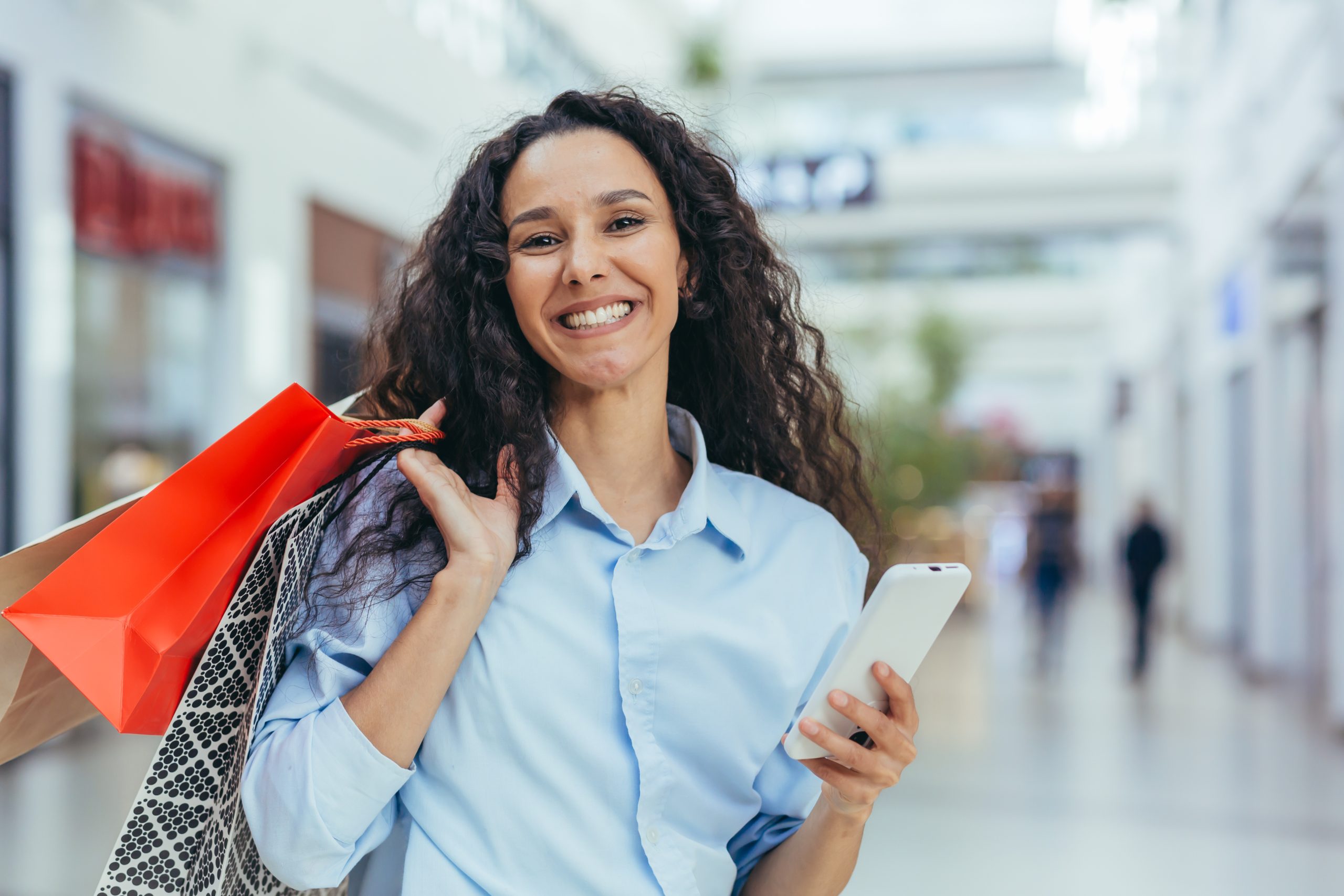 imagem blogpost Capital de giro. Mulher sorrindo, segurando sacolas de compras e um celular em um ambiente comercial.