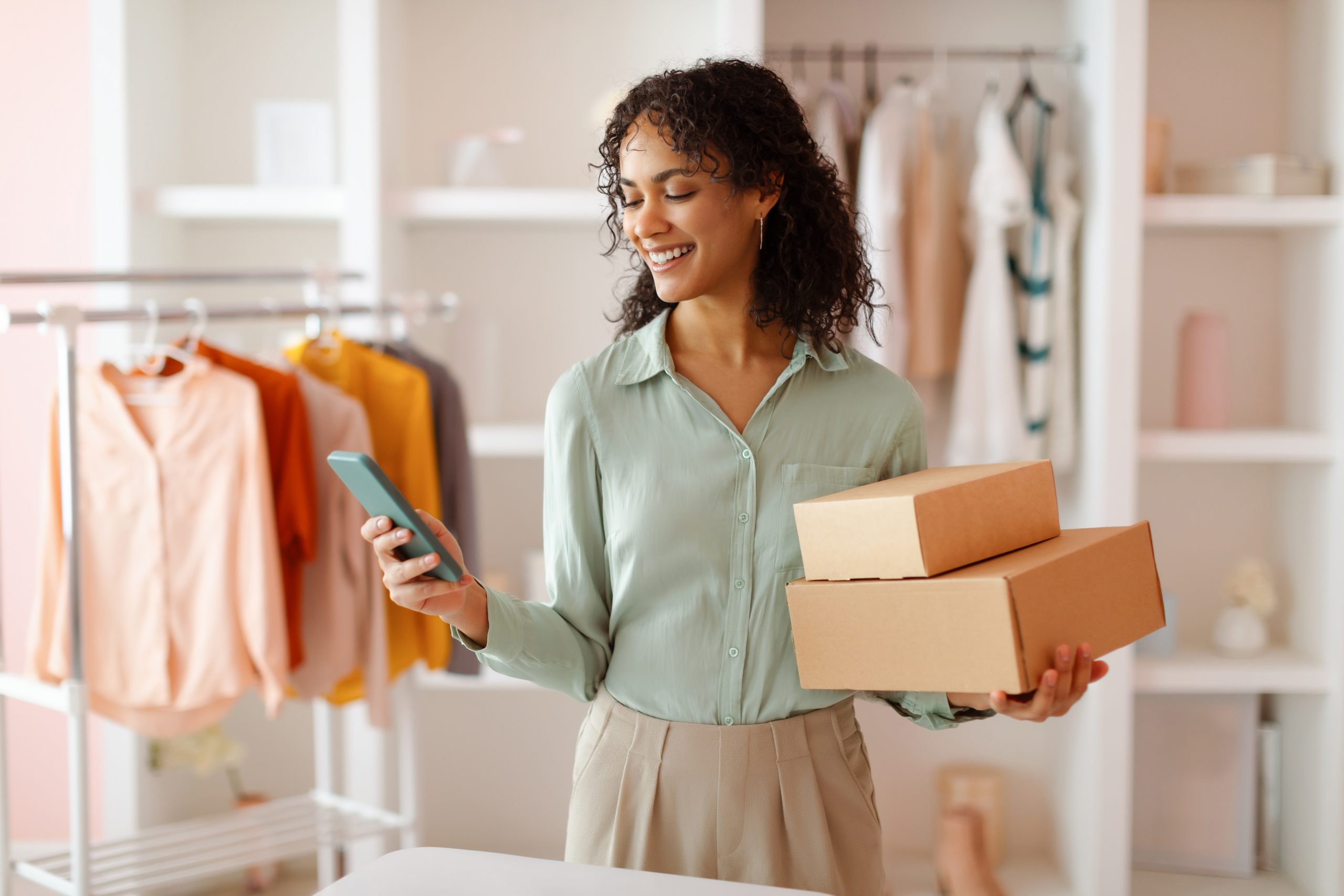 imagem blogpost MEI final de ano. Mulher sorrindo, segurando caixas de encomendas e utilizando o celular em uma loja de roupas.